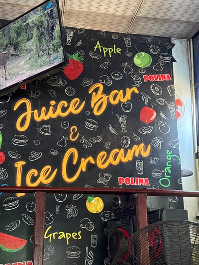 The image shows a black wall with colorful drawings of fruits, ice cream, and food items. Text in yellow and green reads "Juice Bar", "Ice Cream", "Apple", "Orange", and "Grapes". There are also drawings of strawberries, apples, oranges, grapes, watermelons, burgers, and ice cream cones. A television screen is mounted on the upper left side of the wall, displaying an image of a deer in a forest. The wall appears to be in a food establishment, possibly a juice bar or cafe. The context suggests this is a location in Walasmulla, Sri Lanka, as indicated by the prompt.
