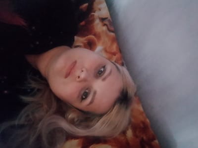The image shows a close-up, tilted portrait of a young woman lying down, with her head angled towards the camera. She has light blonde hair with dark roots and is wearing a dark, possibly black, top with some faint lighter colored circular patterns. Her face is illuminated from the front, highlighting her fair skin, visible right eye with eyeliner, and slightly parted lips. She is lying on a surface with a patterned fabric, featuring warm tones of orange, brown, and cream, resembling animal fur or a similar textured print. The background consists of a light blue-grey surface, possibly a wall or a bedsheet, which is out of focus. The overall lighting is soft and appears to be from an indoor source. There is no indication of the specific location within Изыхские Копи, Russia, as the image focuses solely on the individual and her immediate surroundings.