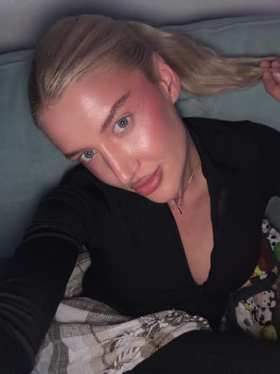 A blonde female is depicted in a close-up, slightly angled shot within an indoor setting. She has light blue eyes and fair skin exhibiting prominent reddish blush across her cheeks and forehead. Her blonde hair is pulled back in a ponytail, with her right hand grasping a portion of it. She is dressed in a dark, long-sleeved garment featuring a V-neckline. A silver-colored chain with a small cross pendant is visible around her neck. Her mouth is closed with a slight pout. A patterned blanket with white fringe covers her lower body and left arm. Behind her, a teal upholstered surface is visible, likely part of a sofa or bed. A colorful, indistinct object is partially visible to her right. The specific location within the city of Yurlovo, Russia, cannot be determined from the visual information provided.