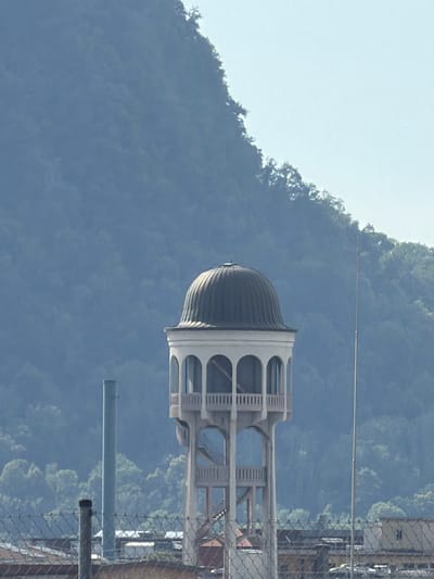 The image, taken from a slightly elevated perspective, captures a striking architectural element set against a backdrop of lush, densely wooded hills. The main subject is a tower-like structure with a prominent, ribbed dome capping its top. Below the dome, the tower features an open, multi-tiered design with arched openings and decorative horizontal banding, suggesting it might be a water tower or a decorative folly. A wire mesh fence runs along the right side of the frame, partially obscuring a glimpse of buildings and possibly a road behind it. In the distance, the rolling hills are covered in a thick canopy of green foliage, with a clear, pale sky above. The lighting suggests a bright day, likely late morning or early afternoon. The scene appears to be an outdoor environment, possibly a park or an area with industrial or civic structures integrated with the natural landscape. No people are visible in the frame, and there is no discernible text.