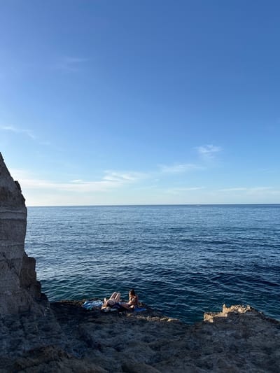 The image depicts a coastal scene in Sperone, Italy, under a clear, bright blue sky with sparse, wispy clouds. An expansive, deep blue sea occupies the mid-ground, meeting a distinct horizon line. Small ripples are visible on the water's surface, and a small, reddish-orange buoy floats in the sea to the left of center. A faint white wake, possibly from a distant vessel, is visible near the horizon.
On the left side of the frame, a large, rugged cliff face, light grey in color and displaying horizontal striations, extends vertically from the bottom edge towards the top. In the foreground, a dark, textured rocky platform extends from the right and foreground towards the cliff and the sea.
Two adult females are situated on a flatter section of this rocky platform, adjacent to the water's edge. Both are wearing swimwear. The female on the left is lying supine on a blue and white striped towel, facing upwards, with her legs slightly bent. The female on the right is seated upright on a blue towel, facing towards the water or slightly towards the first individual. Dark footwear is visible near her feet on the towel. Both individuals appear to be relaxing and sunbathing. The scene is brightly illuminated by natural light, casting subtle shadows on the rocky formations.