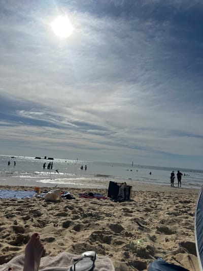 A beach scene under a bright, partly cloudy sky with the sun low on the horizon. People are seen at the water's edge, some silhouetted figures are in the distance. The foreground displays sandy beach, a person's foot, sunglasses, and personal belongings including clothes and bags. A section of a beach tent is visible at the upper edge.