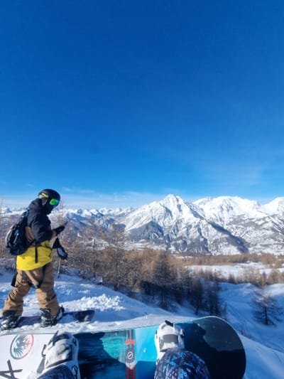 The image displays a snowy mountain landscape under a clear blue sky, situated in the area of Cesana Torinese, Italy. In the immediate foreground, the perspective is from a person's point of view, showing two snowboard boots strapped into a blue, black, and white snowboard. In the midground, a second individual stands on a snowboard, facing slightly left. This person wears a dark helmet with bright green goggles, a yellow and black jacket, light brown pants, and a dark backpack. They are actively holding a dark smartphone in their gloved hands. The terrain consists of fresh white snow, with scattered bare-branched deciduous trees visible between the midground and the distant mountains. The background features a panoramic expanse of multiple rugged, snow-covered mountain peaks under a clear, brilliant blue sky.