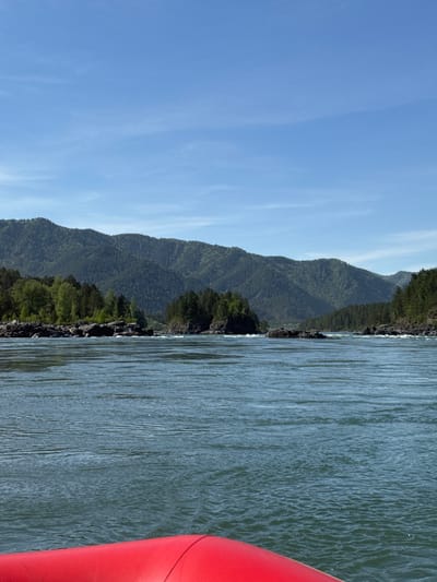 The image depicts a landscape with water, mountains, and sky. The foreground shows part of a body of water with visible surface ripples. To the left of the water are mountains covered in green vegetation. The sky is blue with some wispy clouds. A red inflatable object is partially visible on the right side of the image, possibly a boat or raft. The location is likely near Ust-Sema, Russia, given the mountainous landscape and presence of a river.