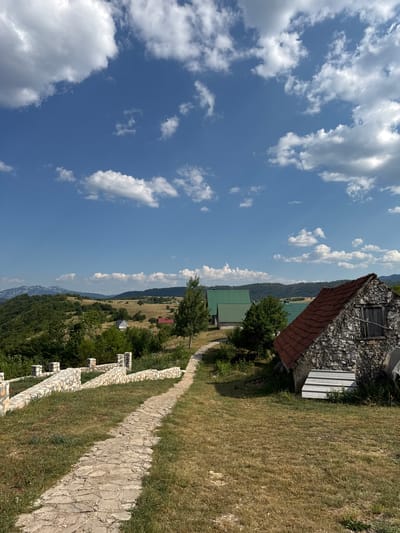 The image depicts a rural landscape with a clear blue sky dotted with white clouds. In the foreground, a stone pathway winds through dry grass. To the left of the path, a stone wall with decorative pillars runs along a raised terrace. On the right side of the path, an old stone building with a red tiled roof stands beside a smaller structure with a white corrugated metal roof. Further in the distance, several modern houses with green roofs are visible among trees. The background reveals rolling hills covered in lush green vegetation, with distant mountains forming a hazy horizon. The scene appears to be set in the village of Rudinice, Montenegro, a location known for its natural beauty and rural charm. There are no people or animals visible in the image, and no actions or interactions are taking place.