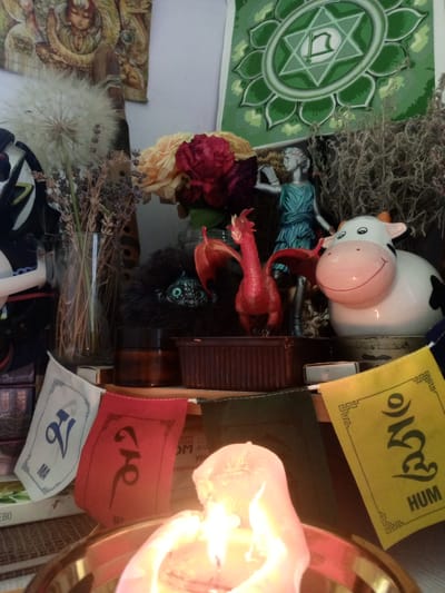 The image depicts an altar or shrine, likely indoors, with a prominent lit candle in the foreground. The altar features a collection of symbolic objects. To the left, a large, dried dandelion seed head stands in a glass vase filled with dried grass and lavender. Behind this, a dark, furry object with large green eyes is partially visible. A small red dragon figurine is positioned in the center, with its wings spread. To the right of the dragon is a white ceramic cow figurine with black spots and a smiling face. Behind the dragon and figurine, a small statue of a person in a blue robe stands. Several prayer flags with Tibetan script are displayed across the front of the altar; one is white with blue lettering, one is red with black lettering, and one is yellow with black lettering and the word "HUM" at the bottom. Behind these, a dark green fabric hangs. A large green mandala-like tapestry is visible at the top left, and another tapestry with a figure is at the top center. The overall impression is of a personal spiritual space. The image does not contain any specific visual cues to indicate its location within Пожарское сельское поселение, Ukraine.