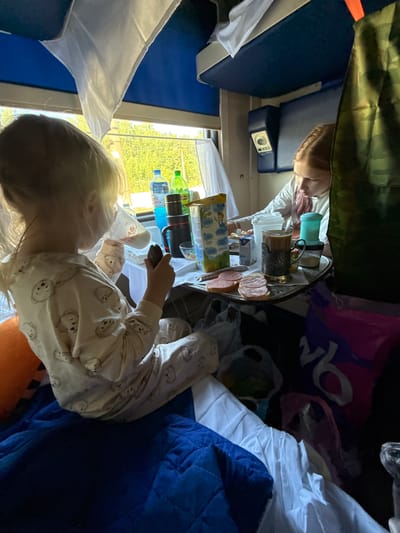 Two young girls are seated inside a train compartment. The girl in the foreground, facing away from the camera, is wearing pajamas with a teddy bear pattern and is holding a cup and a piece of food. She is seated on a blue quilted blanket. The second girl, with red hair, is seated opposite her, looking down at a tray of food and drinks. On the tray are sandwiches with sausage, a cup of coffee, a sugar dispenser, and other items. The compartment appears to be equipped with sleeping berths above the seats, and there are bags and luggage visible in the background. The environment suggests a train journey, possibly in Russia, given the mention of Stupino, Russia. The lighting suggests it is daytime, with sunlight coming through the window, illuminating the trees outside.