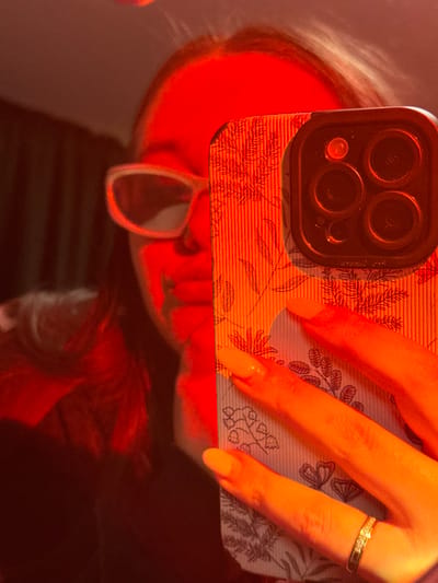 A person is depicted in a close-up, reflected or self-captured image, under strong red-orange illumination. The individual, primarily visible as a partial reflection or through a selfie perspective, is wearing light-framed sunglasses. Their right hand (viewer's left) holds a smartphone in the foreground, obscuring the lower half of their face. The smartphone features a case with a textured, ribbed surface and a botanical pattern of leaves and lily-of-the-valley motifs. The phone's multi-lens camera array is visible in the upper right quadrant of the image. The hand holding the phone has bright orange painted fingernails, and a metallic, possibly gem-encrusted, band ring is worn on the ring finger. The background is dark and out of focus, suggesting an indoor setting. This scene occurs in Prikubanskiy, Russia.