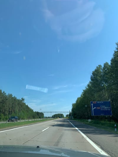 The image depicts a view from a vehicle traveling on a highway in a rural area. The sky is clear blue with some wispy clouds. On the right side of the road, a blue highway sign indicates directions. The top part of the sign displays "P23 МІНСК" with an arrow pointing straight ahead, signifying Minsk as a destination via route P23. Below this, it shows "УЗДА" and "P68 НЕГАРЭЛАЕ ПУХАВІЧЫ" with an arrow pointing to the left, indicating Uzda and directions to Negarelaya and Pukhavichy via route P68. At the bottom of the sign, "300M" is visible. The highway has multiple lanes, and trees line both sides of the road. A dark SUV is visible in the distance on the left lane. The overall scene suggests a sunny day on a road trip in Belarus, specifically within the vicinity of Гацукоўскі сельскі Савет.