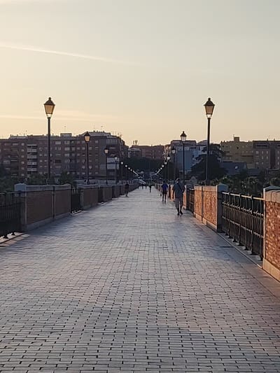The image depicts a view of the Puente de Palmas in Badajoz, Spain, during what appears to be early morning or late evening due to the soft, warm lighting. The bridge, constructed with a cobblestone pathway, is lined with traditional lampposts on both sides. Several individuals are walking along the bridge, some in the distance and one closer to the foreground, a man with a striped shirt and shorts walking away from the camera. The background features a city skyline with multi-story buildings and some greenery. The sky is a pale yellow and orange, suggesting a sunrise or sunset.