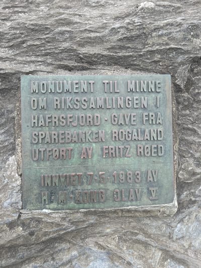 This is a close-up, high-angle, eye-level shot of a bronze plaque affixed to a rugged, gray rock face. The plaque is vertically oriented and appears to be weathered, with a green patina. The text on the plaque is raised and reads: "MONUMENT TIL MINNE OM RIKSSAMLINGENI HAFRSFJORD GAVE FRA SPAREBANKEN ROGALAND UTFØRT AV FRITZ ROED INNVIET 7.5.1983 AV H.M. KONG OLAV V". The text indicates that the monument commemorates the national gathering in Hafrsfjord, was a gift from Sparebanken Rogaland, executed by Fritz Roed, and inaugurated on May 7, 1983, by His Majesty King Olav V. The rock behind the plaque is rough and textured, suggesting an outdoor setting, possibly a historical or memorial site. The lighting is even, indicating it is daytime, and there are no strong shadows. The image is likely taken in Stavanger, Norway, given the mention of Hafrsfjord.