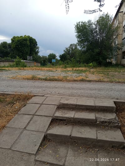 A daytime exterior shot shows a slightly overgrown outdoor area. In the foreground, a set of uneven concrete steps with square textured tiles is visible. Beyond the steps, a gravel path leads to a grassy area with some dry patches. In the background, several trees are scattered throughout the scene. A tall building with balconies stands on the right side of the image. A basketball hoop and backboard are visible in the distance, and a second basketball hoop is present further back. The sky is overcast, and a bird is in flight. The timestamp "22.06.2026 17:47" is overlaid on the bottom right corner of the image.