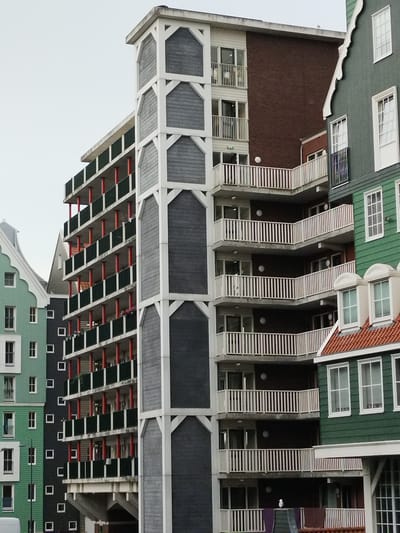 The image shows a close-up, low-angle shot of the Inntel Hotel Zaandam, a contemporary hotel located in Zaandam, Netherlands, notable for its façade composed of 70 traditional Zaan houses. The hotel's exterior features a stacked, fragmented architectural style, with various building sections, colors, and balcony designs visible. Prominently displayed is a light-colored, multi-story tower with white diagonal bracing against a dark grey textured material. To the left, a teal-colored building with white-trimmed windows and gables is partially visible, a characteristic architectural element of the Zaan region. Adjacent to the tower is a section of the hotel with staggered balconies featuring white railings. Further to the right, the green façade of another part of the hotel, also with white-trimmed windows and gables, is visible. Several balconies are visible, some with laundry drying, and a seagull is perched on one of the railings. The sky is overcast, suggesting a cloudy day. The hotel's unique design integrates historical Dutch architectural motifs with modern construction.