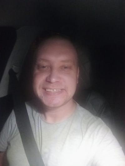 A man with fair skin, a bald head, and stubble is smiling at the camera while wearing a light gray t-shirt and a seatbelt. He is seated in a vehicle, indicated by the seatbelt and the interior of the car visible behind him. The lighting suggests it is nighttime or a dark environment. No specific details about the location within Кадзінскі сельскі Савет, Belarus are discernible from the image.