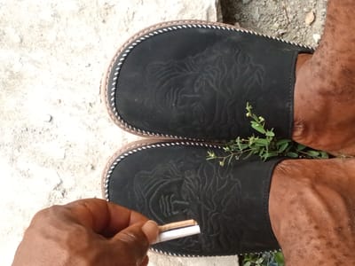 The image shows a person's feet wearing black open-toed slippers with a decorative embossed pattern of what appears to be a lion's head. The slippers have a light-colored trim around the edges with white stitching. The person's skin is brown. In the foreground, a hand is holding a partially rolled cigarette. The background consists of a light-colored, textured surface that could be concrete or sand, and some dry dirt with scattered pebbles and a few small green plants. The scene appears to be outdoors, as suggested by the natural ground cover and lighting. The image is a close-up shot, focusing on the feet, slippers, and hand. The time of day and weather are not explicitly indicated, but the lighting suggests it could be daytime. The location context is Dongo, Nigeria.