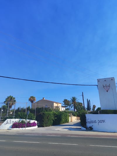 The image depicts a sunny day in Els Poblets, Spain, with a clear blue sky and visible power lines. In the foreground, a paved road runs horizontally. To the left of the road, a white, curved structure with the text "Almadrava park" written in cursive is adorned with vibrant purple flowers. Behind this, a chain-link fence encloses an area with palm trees and dense green bushes. A light-colored building with a tiled roof is visible behind the vegetation. On the right side of the image, a tall, white, rectangular structure, possibly an electrical utility box, stands near the road. It has a red, shield-shaped emblem with some sort of design at the top and electrical components attached. More palm trees and greenery are visible on this side as well, with a portion of a white wall displaying the words "Almadrava park" in black cursive. The overall scene suggests an entrance or boundary to a park or residential area.