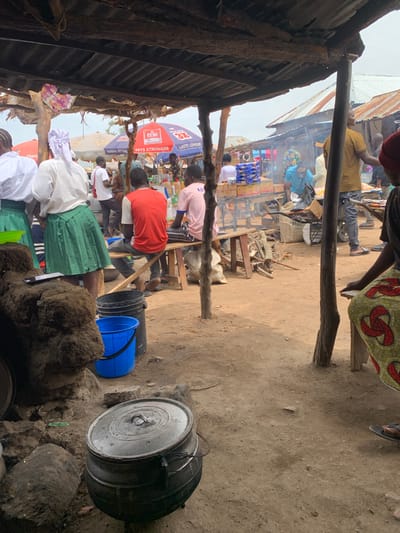 This image depicts a busy market scene in Dull A, Nigeria. Several people, dressed in various casual attire, are visible. Some are seated, others appear to be standing and interacting with each other or the goods on display. The setting is an open-air market with makeshift stalls constructed from corrugated metal and wooden planks. In the foreground, a large, dark pot sits on the ground, suggesting food preparation is underway. Several buckets, one blue and one black, are also present. In the background, more stalls are visible, filled with various items, including what appear to be bottles of liquids and packaged goods.

Smoke is rising from a fire in one of the stalls, indicating cooking is actively happening. The ground is dusty and uneven, typical of an outdoor market. The lighting suggests it is daytime, possibly late morning or afternoon, with no strong shadows indicating harsh sunlight. The overall atmosphere appears active and bustling, with a sense of everyday commerce.

Visible text includes branding on a red and blue umbrella in the background, partially obscured but showing "SETTS STRONGER" and "LASTS LONGER". Another partially visible sign says "Coca-Cola". The scene conveys a sense of community and daily life in a Nigerian market.