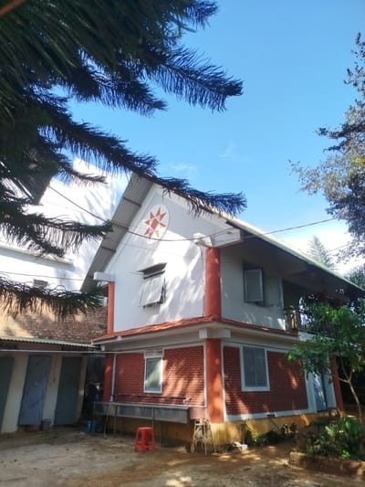 The image depicts a two-story building with a red brick base and white upper level, located in An Phu Ward, Vietnam. The building has a pitched roof with a red and white star design on the gable. Windows are visible on both levels. A covered porch or balcony extends along one side of the upper level.
In the foreground, the ground is dirt with patches of vegetation. A red plastic stool sits near a metal trough or sink. Adjacent to the main building is a smaller structure with blue doors. Overhanging branches from trees obscure parts of the sky and building. The sky is blue with scattered clouds.