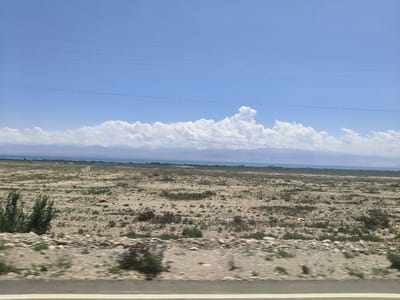 The image depicts a wide, arid landscape under a bright, blue sky with scattered white clouds. In the foreground, there is a road with a white line, suggesting a moving vehicle took the photo. The terrain is mostly dry, with sparse vegetation consisting of low shrubs and rocks. In the mid-ground, a body of water, possibly a lake or sea, is visible, bordered by a distant, hazy mountain range capped with snow. The overall impression is one of a vast, natural setting. The time of day appears to be midday, given the clear sky and bright sunlight. The weather seems fair and sunny, typical of a summer day. The location is identified as Toru-Aigyr, Kyrgyzstan, which aligns with the arid landscape and mountainous backdrop. There are no visible people or objects other than the natural elements and the road. The scene evokes a sense of remoteness and natural beauty. There is no visible text in the image.
