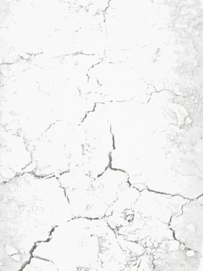The image displays a close-up, top-down view of dry, cracked earth, resembling a parched ground surface. The texture is rough and uneven, with intricate patterns of fissures and crevices running across the frame. The color palette is predominantly monochromatic, consisting of various shades of white and grey, with some darker shadows accentuating the depth of the cracks. The lighting appears bright, possibly indicating an outdoor scene under daylight conditions, contributing to the bleached appearance of the material. There are no discernible human figures, animals, or man-made objects present in the image. The overall impression is one of dryness and desiccation, suggesting an arid or weathered environment. There is no information available within the image or its context to place this scene in Matchino, Russia, or any specific geographical location.