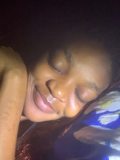 A close-up captures a person with closed eyes and a serene expression. Their face is illuminated, revealing smooth, glowing skin and full lips curved into a gentle smile. They are resting their head on their arm. A pillow with a blue and white floral pattern is near their head. The lighting creates a soft shadow on one side of their face.