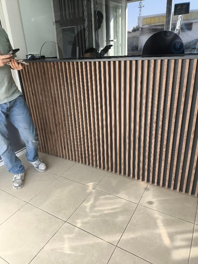 A partially visible person in jeans and a green shirt stands beside a wooden reception desk with vertical slats. The desk has a dark counter and a transparent barrier. Behind the desk, a black dome-shaped object with a currency symbol is visible. Sunlight streams in from the right, illuminating the tiled floor. The scene is indoors with a glimpse of an exterior building seen through the glass behind the desk.