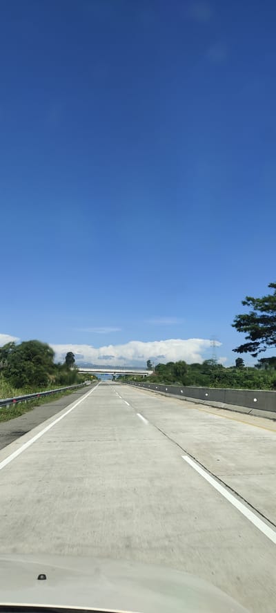 The image captures a view from a vehicle traveling on a highway, likely in Lonbaroh, Indonesia, given the context. The road is made of concrete with clear white lane markings. The sky is a vibrant, cloudless blue, occupying the majority of the upper frame. In the distance, a bridge spans across the highway, and beyond that, lush green trees and hills are visible, with a few scattered clouds. The foreground shows the hood of the vehicle, indicating the perspective is from the driver's or passenger's seat. The overall scene depicts a clear, sunny day on a well-maintained highway, surrounded by natural greenery.