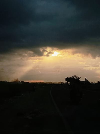 The image depicts a dramatic sunset scene in Keude Ulim, Indonesia. Dark, ominous clouds dominate the upper portion of the sky, with a sliver of bright, golden sunlight breaking through the center, casting visible rays downwards. The horizon is lined with silhouetted trees and structures, with faint smoke plumes suggesting industrial activity or fires in the distance. In the foreground, a dark, asphalt road curves to the left. A silhouette of a person riding a motorcycle is visible on the right side of the road, carrying a large bundle of what appears to be vegetation on the back. Another motorcycle is faintly visible further down the road. The overall atmosphere is one of dusk settling over a rural or semi-industrial landscape.