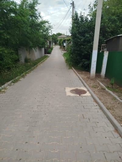 The image depicts a paved pathway in a village setting, likely in Ceadîr-Lunga, Moldova. The pathway, made of interlocking gray pavers, extends into the distance, flanked by lush green trees and vegetation on the left and a green metal fence and more trees on the right. A concrete curb runs along the right side of the pathway. In the foreground, a manhole cover is visible embedded in the pavement, surrounded by a square concrete frame. Utility poles and power lines are present in the background, along with houses and other structures characteristic of a rural or semi-urban environment. The sky is overcast, suggesting a cloudy day. There are no people visible in the image.