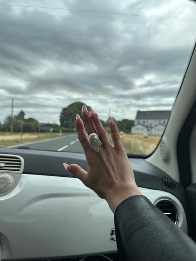 An overhead shot shows a woman's left hand with long, light-pink nails and a large, irregular pearl ring on her index finger as she waves outside a car window. The background is blurred, showing a country road with fields and buildings. The interior of the car, with its white dashboard and black leather accents, is visible in the foreground. The sky is overcast with gray clouds.