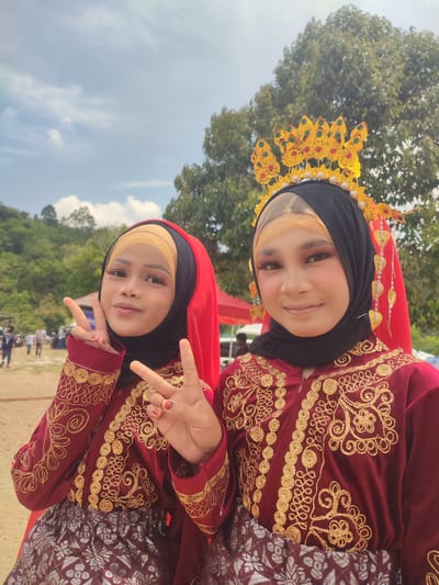 Two young women, adorned in traditional attire, pose for a photograph outdoors. Both are wearing matching maroon velvet tops with intricate gold embroidery and black hijabs. The woman on the left has a yellow hijab underneath, while the woman on the right wears a golden headpiece adorned with gemstones. The woman on the left is making a peace sign with her right hand and her left hand is out of frame. The woman on the right has her hands also out of frame. They are standing in a dusty, open area with a few people and tents visible in the background. Lush green trees and a hilly landscape under a cloudy blue sky complete the scene, suggesting a location in Pantonrayeuk Satu, Indonesia.