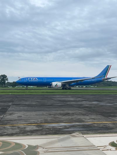 The image shows an ITA Airways airplane parked on an airport tarmac. The plane is primarily blue with ITA Airways branding. The tail fin features the Italian flag colors. The ground around the plane consists of concrete and grass. The sky is cloudy. Based on the location information, this scene is likely at Malabo International Airport in Malabo, Equatorial Guinea.