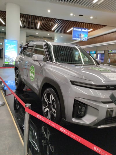 The image shows a gray electric SUV on display indoors. The vehicle has a green circular emblem on its side that depicts a car and a charging station. A red ribbon barrier with the text "KZV Havalimanları" and "KZV Airports" separates the vehicle from the viewer. In the background, there are signs and architectural elements, including wooden slatted ceilings and lights. One sign in the background reads "Depreme Dayanıklı Yapıların Güvenlisi". Another sign on the left side of the image displays the text "Güneş Enerjili Santralı ile yatırım evde başlar". The location appears to be an airport terminal or a public exhibition space. Given the presence of the text "KZV Havalimanları" which translates to KZV Airports, and the text "Güneş Enerjili Santralı ile yatırım evde başlar" which relates to solar energy, this exhibit might be at the Tarsus-Adana-Mersin Airport (TCDD), which serves the Tarsus region in Turkey.
