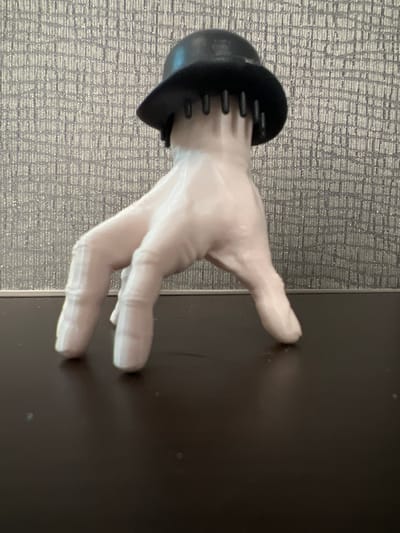 A close-up, medium shot captures a novelty item resembling a disembodied hand wearing a black bowler hat. The hand is pale and stylized, with its fingers bent and extended as if walking. The hat is perched atop the hand, with several dark, thin protrusions emerging from beneath its brim, resembling short hair or bristles. The background is a textured, grey wall with a repeating geometric pattern. The hand rests on a dark, reflective surface, likely a tabletop. The lighting is direct, casting subtle shadows on the surface. No specific location in Columbia, United States of America is discernible from the image.