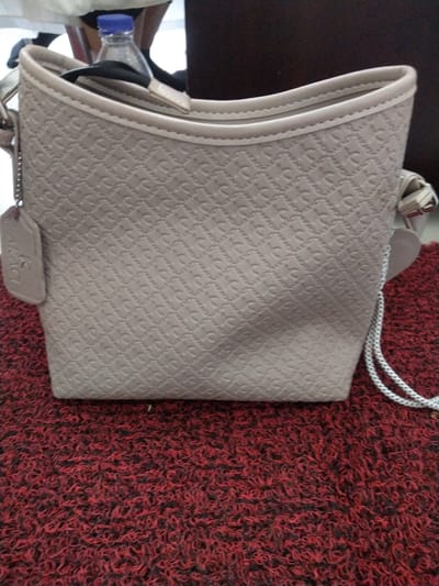 A light tan, textured handbag with a patterned exterior sits on a red, shag-like rug. A clear plastic water bottle with a blue cap is visible inside the top opening of the bag. A small, rectangular tan tag hangs from a silver chain on the left side of the bag. The bag has two thin, light tan straps and a white cord with a black stripe is attached to the right side, looping down and out of frame. The background is dark and out of focus. This image was taken in Pulorayeuk, Indonesia.