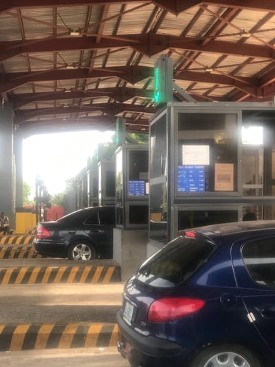 The image captures a bustling toll plaza in Kubare, Nigeria, during bright daylight. Vehicles are actively passing through multiple lanes, with a green arrow illuminated on a traffic light indicating an open lane.
In the foreground, a dark blue car, possibly a minivan or SUV, is prominently featured, with its rear and side visible as it proceeds through a toll lane. Another dark vehicle can be seen further down an adjacent lane. To the left of the vehicle lanes are several glass-enclosed toll booths. One booth clearly displays a "TOLL PLAZA-TARIFF" sign on its window. The overhead structure is a large, covered area supported by reddish-brown metal beams and corrugated metal siding, extending across the toll lanes. The ground features distinctive yellow and black striped markings.
Notable visible text from the toll tariff sign includes:
* "TOLL PLAZA-TARIFF"
* "CARS N 500"
* "SUV N 800"
* "BUSES N 1,000"
* "TRUCKS N 1,600"
* "COMMERCIAL VEHICLES N 500"
A partially visible sign above the tariff also shows "NGEO". The "N" currency symbol confirms the location as Nigeria.