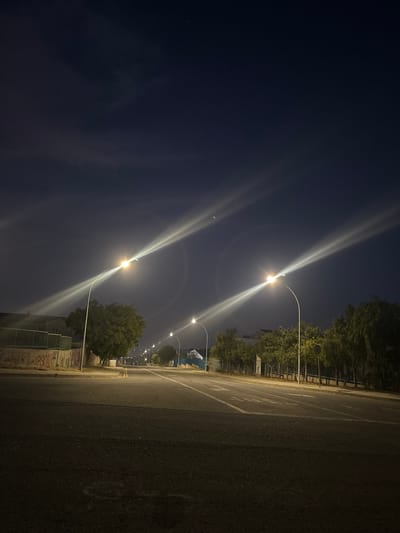 The image shows a street at night, presumably in the Ypsonas Municipality, Cyprus. Streetlights illuminate the area, casting bright beams into the dark sky. Trees line the side of the road. A concrete wall with graffiti is visible. The asphalt road surface is dark.