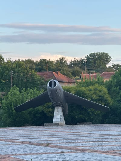The image shows an outdoor monument of a jet airplane in the city of Kamenets, Bulgaria. The airplane is dark gray and mounted on a white pedestal. Its wings are extended, and the nose faces the viewer, revealing the intake. The background consists of lush green trees, red-tiled roofs of houses, and telephone poles with wires. The sky above is a soft blue with wispy clouds. The foreground is paved with rectangular stones arranged in a pattern. The scene appears to be captured during daylight, possibly late afternoon, given the lighting and sky conditions. There are no people visible in the image.