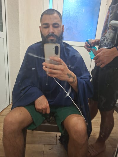 A bearded man in Tarnak, Bulgaria, wearing a blue barber cape and green shorts, is reflected in a mirror while taking a selfie with a smartphone. Another person is partially visible in the reflection, holding a spray bottle. Hair clippings are visible on the floor. The floor is tiled with wood-look planks.