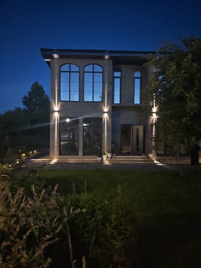 The image shows a modern, two-story house at dusk or night in Надежда Снт, Russia. The house has a gray, stucco exterior with large, dark-framed windows. The upper floor features three arched windows, while the lower floor has a large glass door and two rectangular windows. Exterior lighting illuminates the facade, with vertical lights above the entrance and ground-level lights along the walkway. The front yard is landscaped with green grass, shrubs, and trees. A pathway leads to the entrance, and several small, glowing lights are visible along the path. The sky is a deep blue, indicating evening. No people are clearly visible in the image.