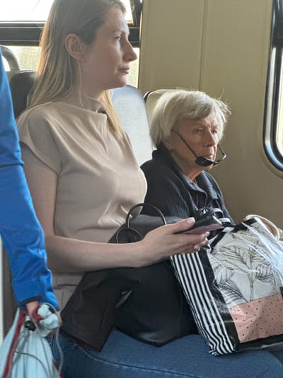The image shows two women seated next to each other on what appears to be public transportation, possibly a train or bus, in Beloostrov, Russia. The woman on the left, with blonde hair, is wearing a tan, short-sleeved top and blue jeans. She is holding a dark-colored smartphone in her right hand and her left arm rests on a black bag. Next to her, to the right, is an older woman with short, gray hair. She is wearing a dark-colored, textured jacket and glasses that are perched on her lower face, near her mouth. She has a bag with a black and white striped pattern and a tropical leaf design on it, partially visible in front of her. The environment appears to be indoors, with neutral-colored walls and seating, and natural light coming from a window on the right side of the frame. The overall scene is casual and mundane, suggesting everyday travel.