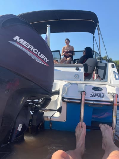 The image depicts the stern of a boat on water, likely on a river or lake, under a clear sky. The lower part of the image is dominated by a large Mercury Four Stroke outboard motor. In the background, people are visible in the boat's cockpit, one appearing to be seated at the helm and another sitting near the front. The boat's ladder is visible, and a person's feet are near the ladder in the water. The boat features blue and white accents. The presence of watercraft suggests the scene could potentially be situated in the city of San Fernando, Argentina, given its location in the Río de la Plata Delta, a popular boating area.