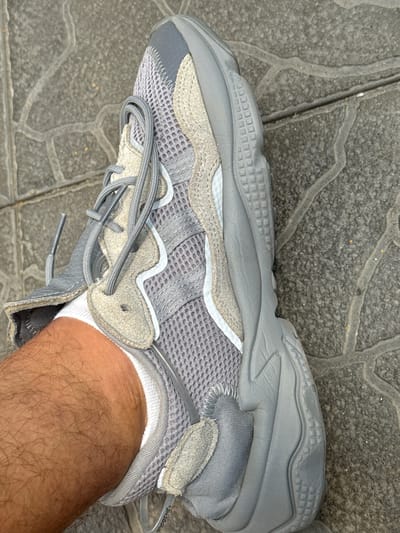 This media file displays a close-up, high-angle shot of a person's left foot wearing a light grey Adidas Yeezy 500 sneaker. The shoe features a mesh upper with suede overlays in various shades of grey and beige. The laces are also grey, and the sole is a thick, sculpted grey material with a patterned texture on the heel and mid-foot. The shoe is resting on a grey tiled surface, with visible grout lines forming an abstract pattern. A portion of the person's lower leg and ankle are visible, covered in dark hair and a white sock. The lighting suggests it is daytime, possibly outdoors. The overall impression is a candid, everyday shot focusing on the footwear and its texture against the textured ground. The setting appears to be urban or a paved outdoor area, consistent with a location context in Suceava, Romania. There is no visible text on the shoe other than a subtle branding on the heel tab which reads "ADI/PUNE".