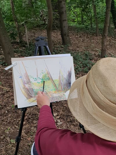 An individual, wearing a burgundy long-sleeved shirt and a wide-brimmed straw hat, is seated in a forest clearing and sketching on a canvas. The canvas, propped on a black tripod easel, is being worked on with a dark pencil. The artist's hand and arm are visible, holding the pencil. The forest floor is covered in fallen leaves and sparse green vegetation. Several trees with textured bark surround the clearing. In the background, another tripod supports a camera or similar device. The overall scene depicts an outdoor artistic endeavor in a wooded environment. The location is identified as a forest in Falkensee, Germany.