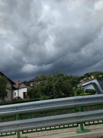 The image depicts a cloudy sky over a residential area in Vyskytná nad Jihlavou, Czechia. In the foreground, a metal guardrail with green supporting structures is visible. Behind the guardrail, several houses with tiled roofs are nestled amongst lush green trees. The sky is filled with dark, heavy clouds, suggesting an impending storm. The overall scene conveys a somber and dramatic atmosphere, characteristic of the approaching bad weather.