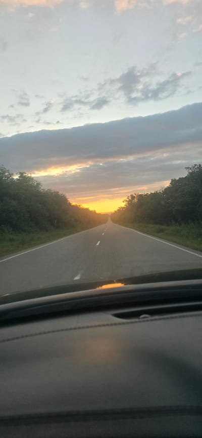 The image depicts a road leading towards a sunset. The sky is filled with clouds, some tinged with orange and yellow hues from the setting sun. A two-lane asphalt road stretches into the distance, bordered by lush green trees on either side. The foreground shows the dashboard of a car, with a reflection of the sunset visible on its surface. The scene suggests a journey through a rural or natural landscape as evening approaches. No specific location in Urozhaynyi, Russia can be identified from the image alone.