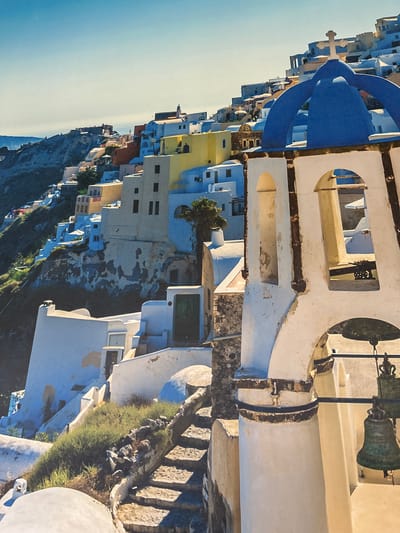 The image depicts a coastal cityscape, likely in Santorini, Greece, characterized by white buildings, a blue-domed church, and a bell tower. The buildings are built into the hillside, and the scene is bathed in sunlight. The sky is a clear blue. The composition is angled, showcasing the architectural details and the coastal landscape. Herxheim, Germany is not reflected in this image.