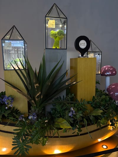 The image depicts a retail display, likely in a shopping mall, featuring decorative elements and merchandise. At the center, a prominent yellow, boat-shaped structure serves as the base for a tiered display. Various glass-enclosed geometric structures, resembling miniature houses or lanterns, are strategically placed on pedestals. One of these houses contains a cartoonish green broccoli character with a smiley face, illuminated from within. Another houses a green leafy plant.

To the right of the broccoli display, a black location marker with "K11" illuminated in white dots stands atop a golden pedestal. Further to the right, two decorative pink mushroom-shaped objects are visible. The entire display is adorned with lush green foliage, including large, spiky leaves and smaller, darker leaves, interspersed with delicate purple flowers. Spotlights illuminate the scene from below, casting a warm glow on the wooden elements and foliage. The background is a plain grey wall. The overall impression is one of artistic and whimsical presentation, possibly for a seasonal promotion or a brand exhibition. The presence of "K11" suggests a connection to a K11 Art Mall, a chain known for its integration of art and retail, often found in major Chinese cities. Without more specific visual cues, pinpointing the exact location within Shenhe District, China, is not possible from the image alone.