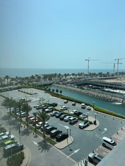 The image depicts a construction site and a parking lot in Ras Al-Khaimah, United Arab Emirates. Several large cranes are visible, indicating ongoing construction. The scene includes a body of water, possibly a canal or the sea, alongside the construction area. Adjacent to the water is a paved parking lot filled with numerous cars, some parked in designated handicap spots. Palm trees line the area, casting shadows on the ground, suggesting a warm, sunny day. The sky is clear and bright blue. The overall impression is one of development and urban expansion in a coastal environment. There is some text visible on the cars and parking spaces, including numbers like "005".