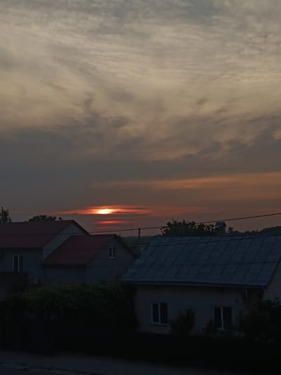 This is a photograph of a sunset over a rural landscape in Pustomyty, Ukraine. The sky is filled with dramatic clouds, tinged with orange and red hues from the setting sun. The sun itself is a bright, small orb peeking through a horizontal band of clouds.
In the foreground, several houses with red and grey roofs are visible. The houses appear to be residential buildings, with windows and some architectural details. Lush green foliage and trees are abundant, obscuring parts of the houses and creating a natural, somewhat overgrown feel to the scene. A power line stretches horizontally across the image, adding a touch of human infrastructure to the otherwise natural setting.
The time of day is clearly sunset, indicated by the low angle of the sun and the warm colors in the sky. The weather appears to be partly cloudy, with a mix of darker, heavier clouds and lighter, wispy ones. The overall mood of the photograph is serene and peaceful, evoking the quiet beauty of a rural evening. There is no visible text in the image.